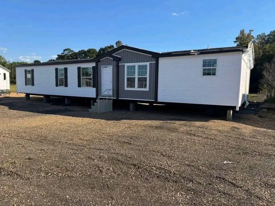 A large, modern manufactured home sits on a gravel lot, featuring white siding with gray accents. Bright blue sky and trees in the background.