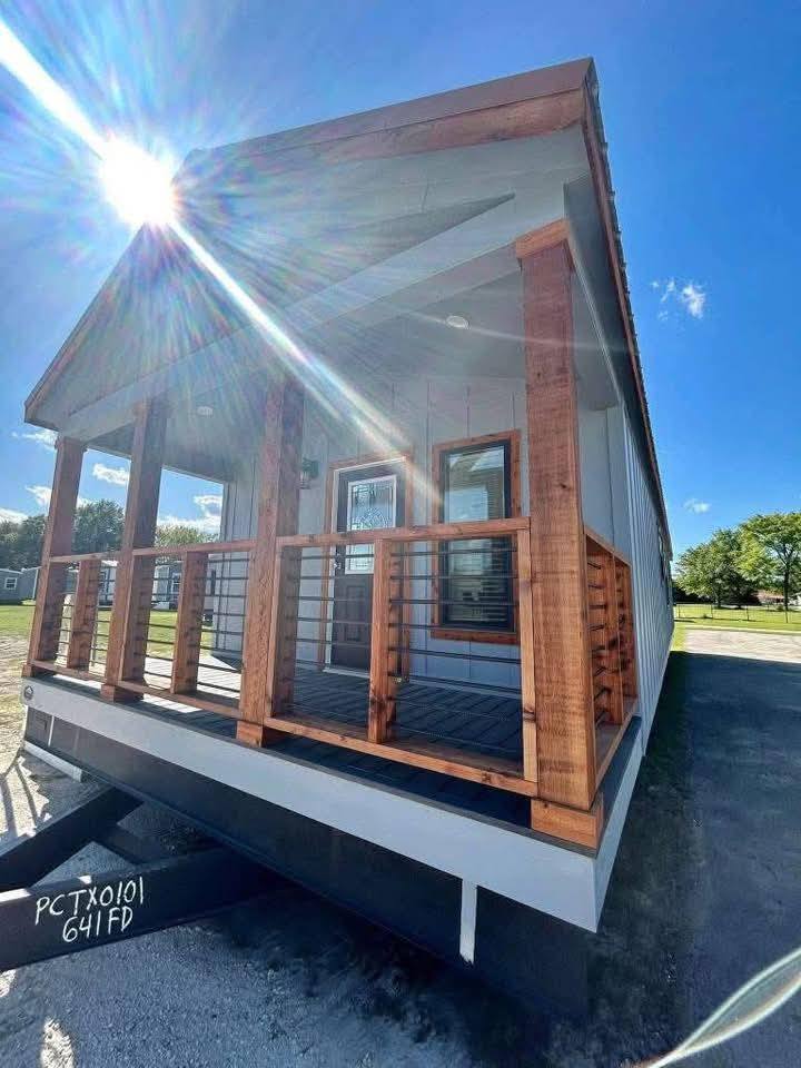 A tiny house with wooden railings and a porch stands under a bright blue sky. Sunlight beams from the top left, casting a warm and inviting glow.