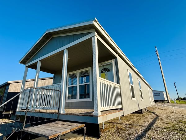 A gray manufactured home with a covered porch and white railing sits on a gravel lot under a clear blue sky, evoking a sense of simplicity and tranquility.