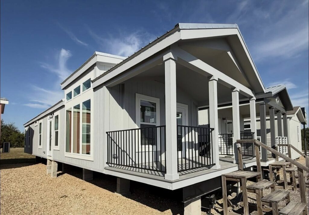 A modern, light-gray manufactured home with a small porch, white trim, and large windows sits on a gravel foundation under a blue sky.