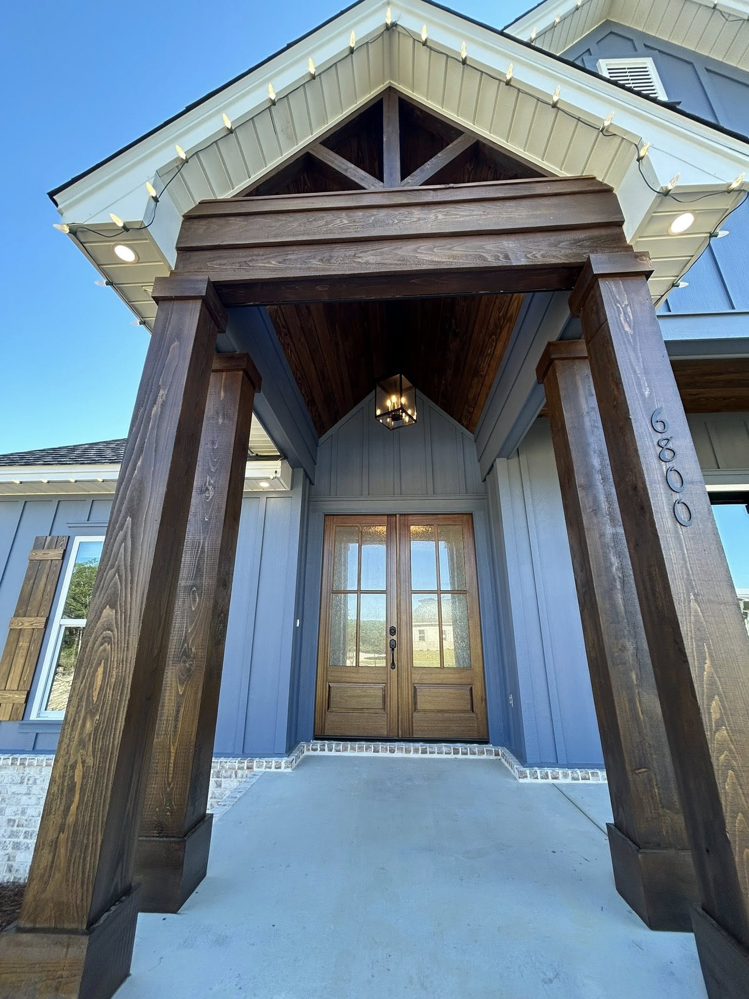 A modern house entrance with a peaked roof and dark wooden columns, featuring a clear glass double door. The blue exterior creates a welcoming vibe.