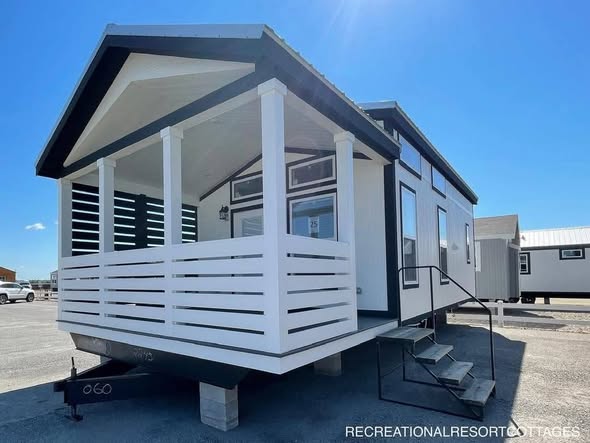 A modern tiny house with white siding and black trim sits on concrete blocks. It has a covered porch, large windows, and stairs leading to the entrance. Bright, clear sky.