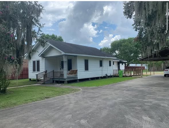A small white house with a black roof sits in a lush yard. A wooden porch is at the front, and a driveway leads up to it. The sky is partly cloudy.