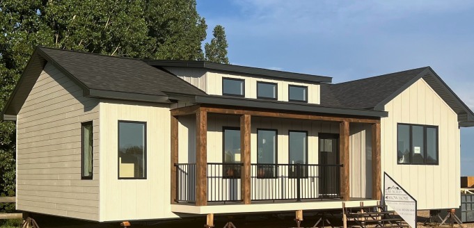 Modern modular home with light siding and dark trim, featuring large windows and a small wooden porch. Surrounded by trees, conveying a sense of tranquility.