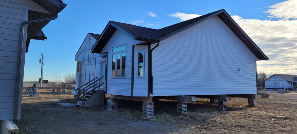 A small white house on stilts stands in a rural area against a clear blue sky. Nearby buildings and barren ground create a remote, peaceful setting.