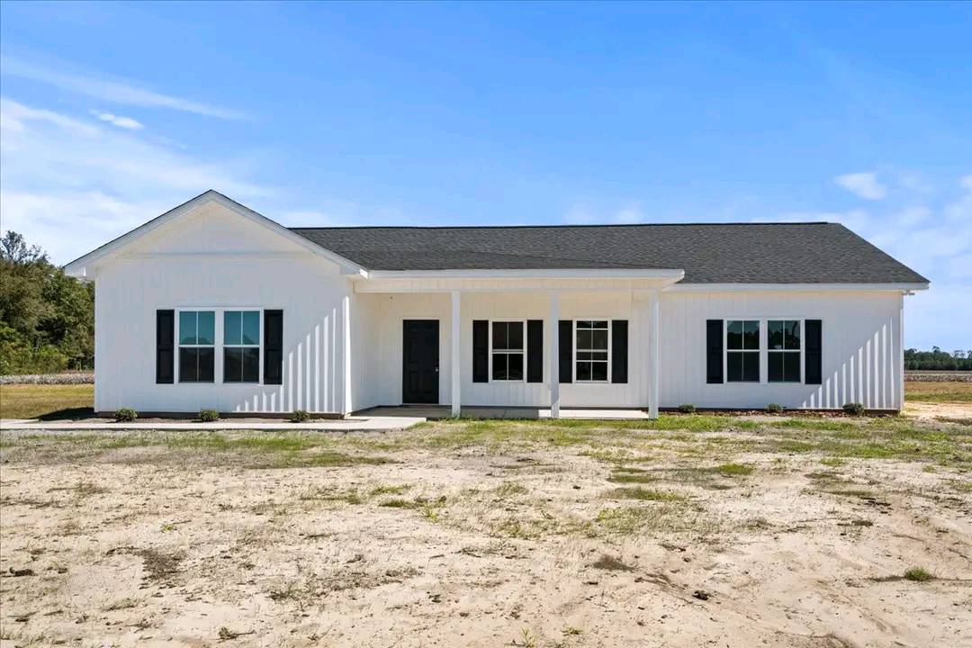 A single-story white house with a black roof and shutters, featuring a front porch with columns. It sits on a sandy, open lot under a clear blue sky.