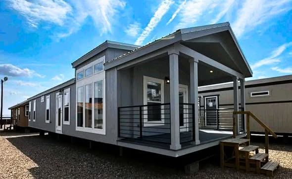 Modern gray mobile home with large windows, a front porch with railings, and wooden steps. Set on gravel under a bright blue sky with clouds.