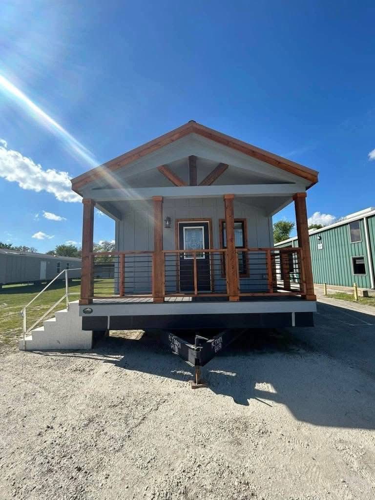 Small, elevated cabin with a wooden porch and peaked roof, set under a clear blue sky with sunlight streaming in. The setting conveys a peaceful, inviting atmosphere.