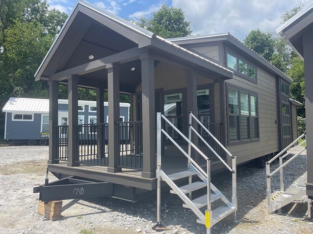 A modern tiny house on wheels with a covered porch and metal railings. The structure sits on gravel, surrounded by trees under a partly cloudy sky.