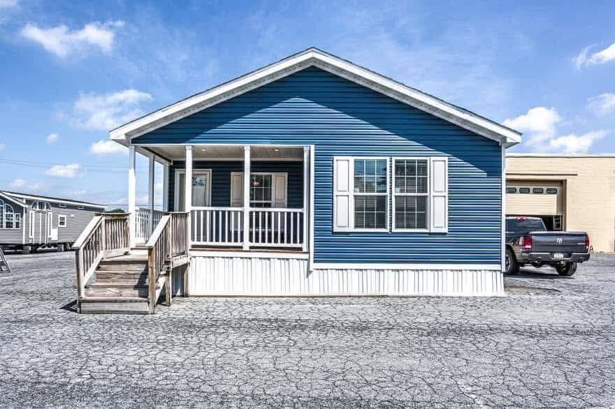 A blue manufactured home with a white porch and steps sits on a cracked asphalt lot. A black truck is parked nearby under a clear blue sky.