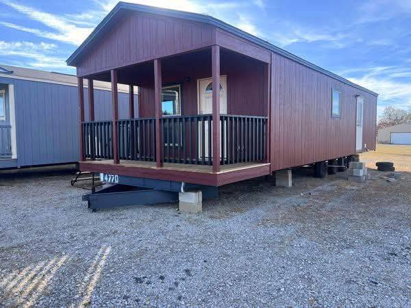 Small, elevated brown cabin with a covered porch and black railings on a gravel lot. Blue sky in the background. Simple and rustic atmosphere.