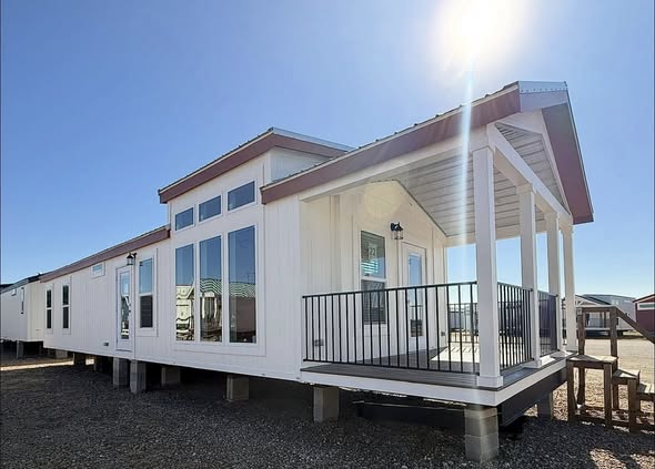 Bright tiny home with large windows and a covered porch, elevated on a gravel area. Sunlight shines overhead, casting a warm, inviting glow.