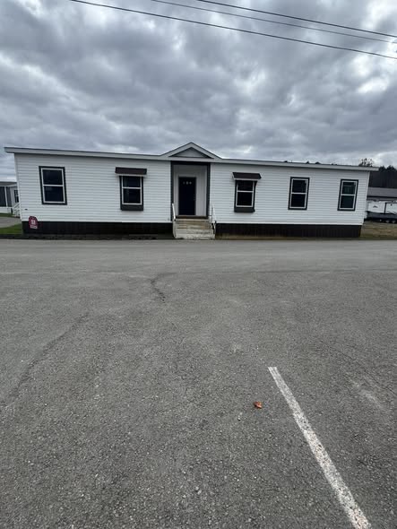 A white modular home with a black foundation sits under a cloudy sky. The building has a central entrance with steps and multiple windows, creating a stark, simple appearance.