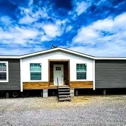 A single-story gray and white mobile home with a centered front door, two large windows, and wooden steps under a bright blue, partly cloudy sky.