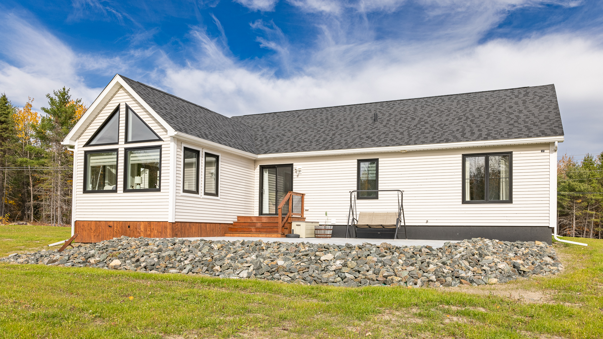 A modern white single-story house with a black roof, large triangular windows, and a porch. It's set against a bright blue sky and surrounded by greenery and rocks.