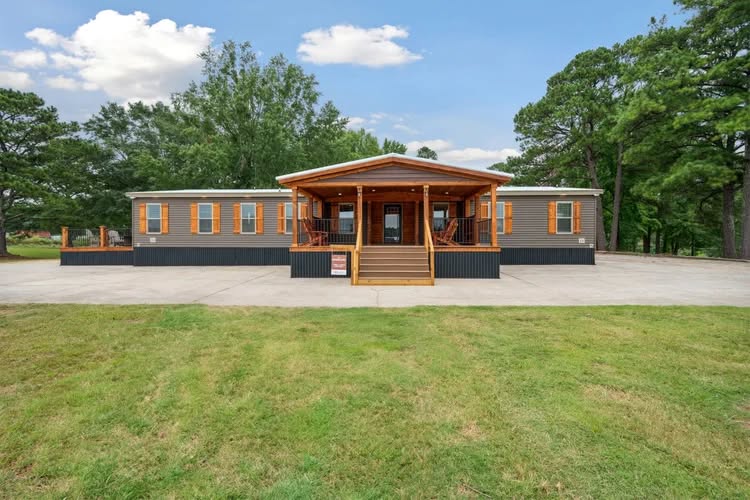 A modern, single-story manufactured home with a wooden porch, surrounded by lush green grass and tall trees, under a clear blue sky.