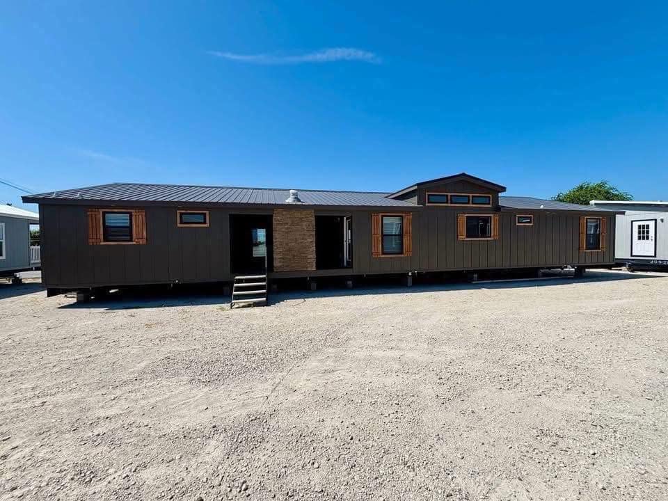 A large modular home with dark siding and wooden trim sits on a gravel lot under a clear blue sky. Steps lead to the entrance, giving a welcoming feel.