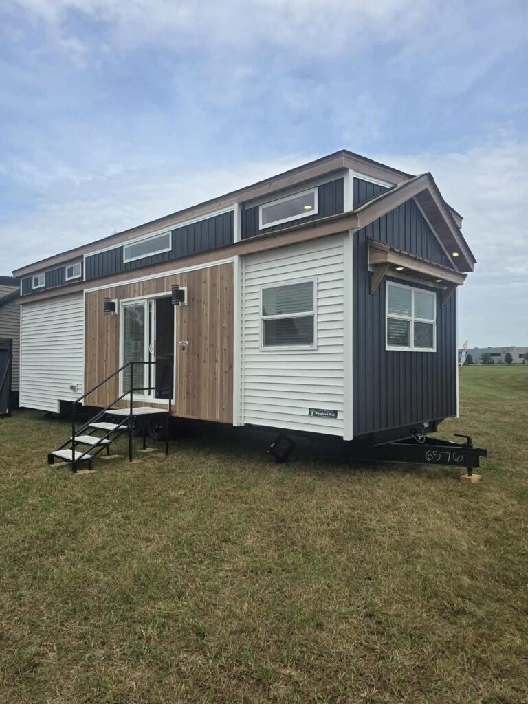 A modern tiny house on wheels with dark blue, white, and wooden exterior stands on a grassy field. It features small windows and a compact staircase.
