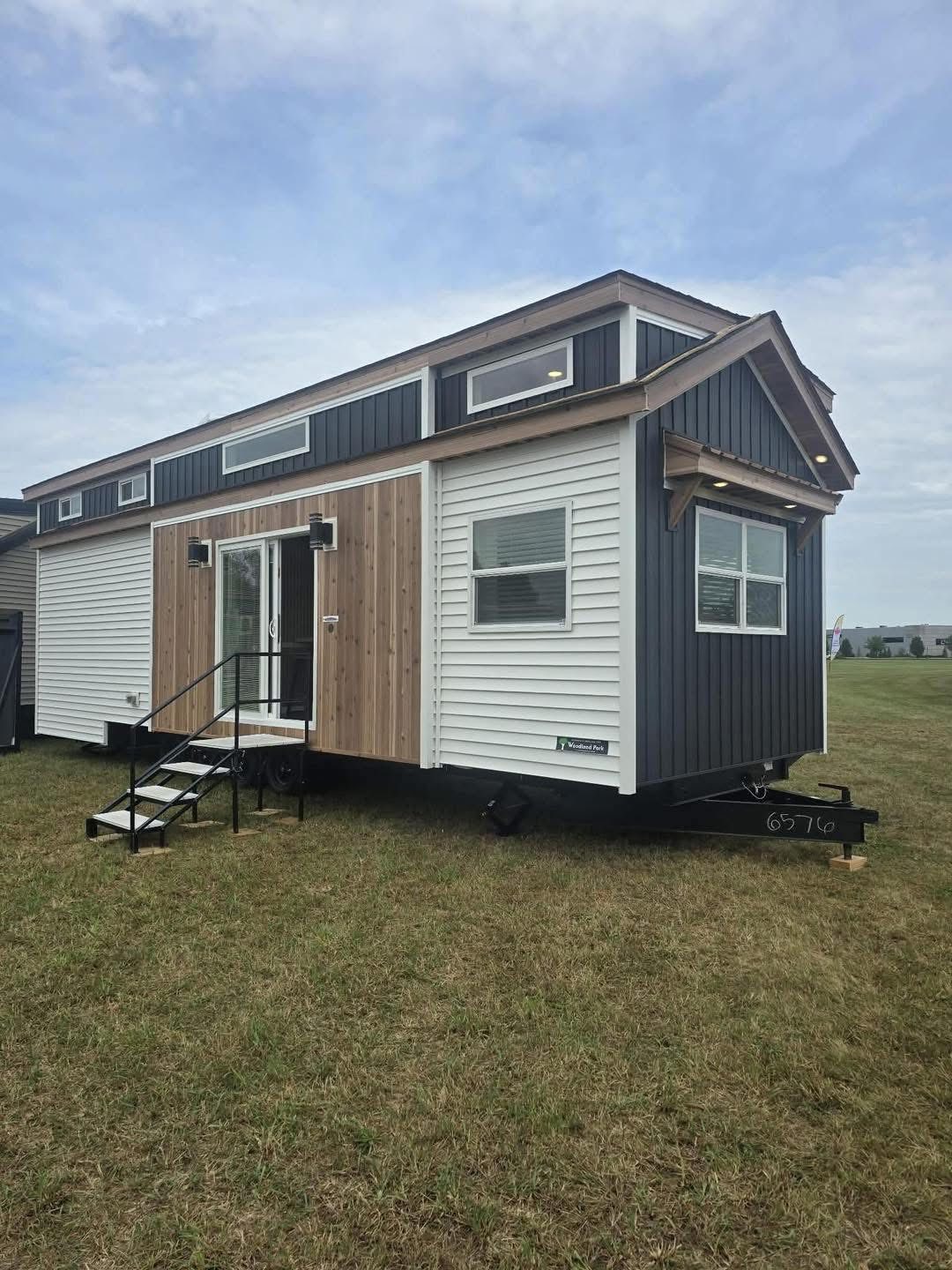 A modern tiny house on wheels with dark blue, white, and wooden exterior stands on a grassy field. It features small windows and a compact staircase.