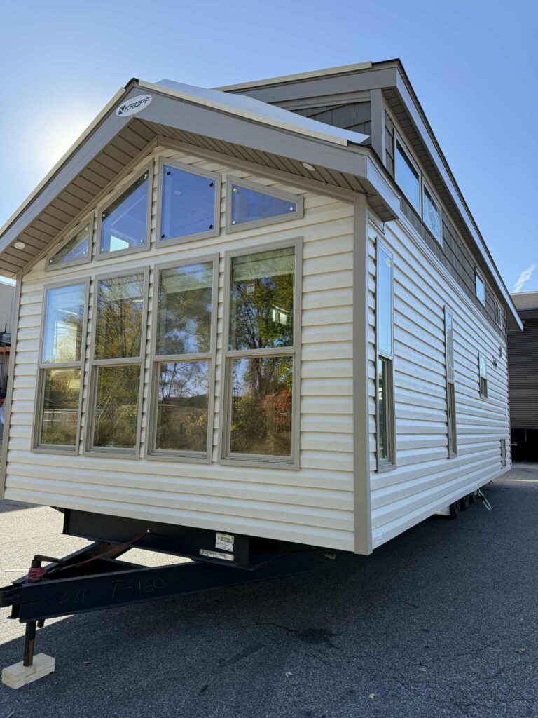A modern mobile tiny house with cream siding and large front windows sits on a trailer. The sky is clear, with sunlight shining above.