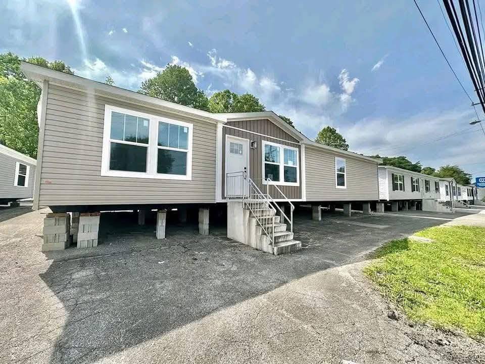 A mobile home on concrete blocks with beige siding and large windows sits in a lot under a partly cloudy sky, evoking a sense of modern simplicity.