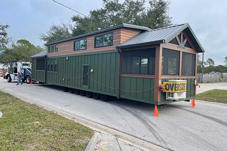 A large green and brown tiny house on wheels marked "OVERSIZE" is being towed down a suburban street, surrounded by trees and vehicles.