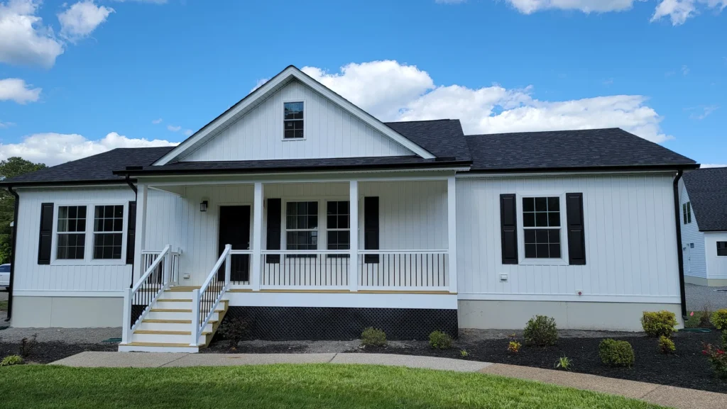 A white, single-story house with a gabled roof under a blue sky. Features include a front porch with railings, black shutters, and a green lawn.