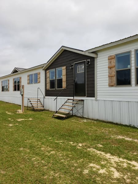 A manufactured home with a dark central section, white siding, and wooden shutters. Two small stairways lead to doors, set on a grassy yard under a cloudy sky.