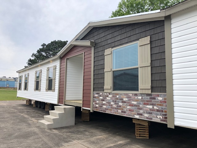 A mobile home with brick and siding exterior sits elevated on blocks. It features large windows with shutters and a small set of concrete steps.