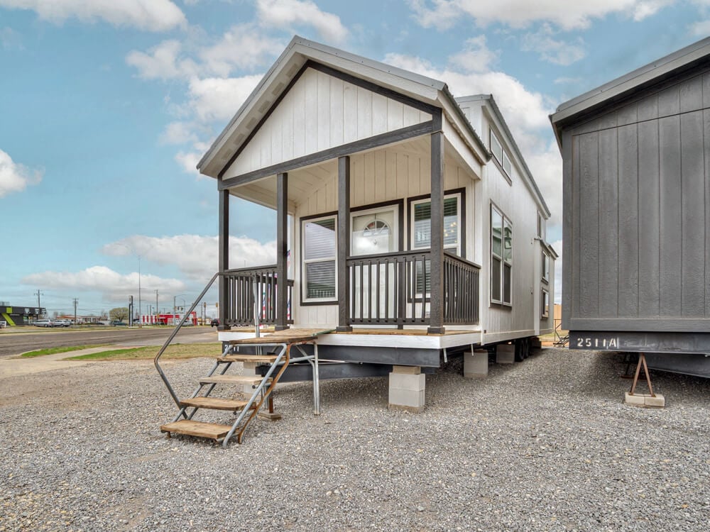 Small modern cabin on raised foundation with front porch, wooden steps, and black railings. Sky is partly cloudy, creating a bright and open atmosphere.