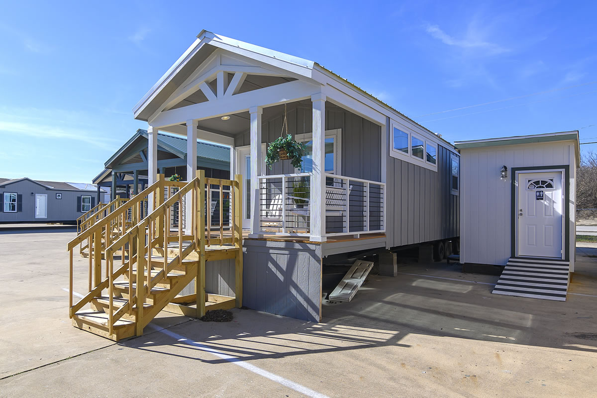 A gray mobile home with white trim, featuring a small porch with wooden steps in sunlight. It has a hanging plant and an adjacent small structure.