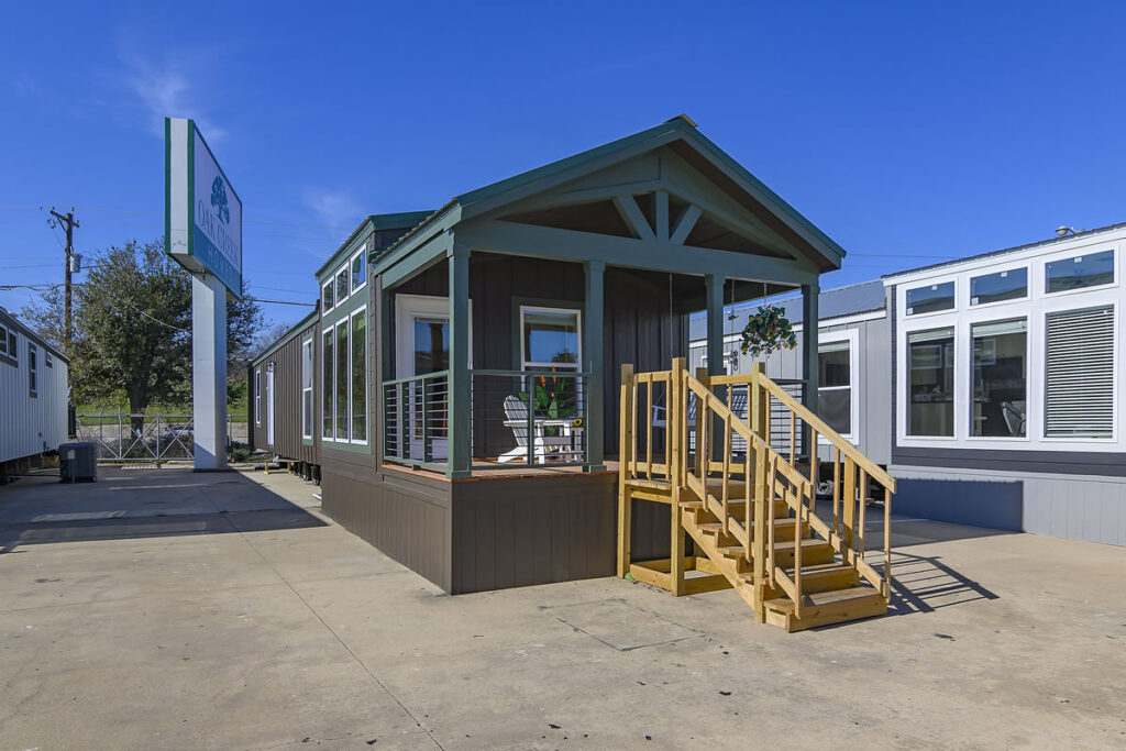 A small, modern modular home with dark siding and a green roof on a sunny day. Wooden steps lead to the porch. Adjacent buildings and blue sky in view.