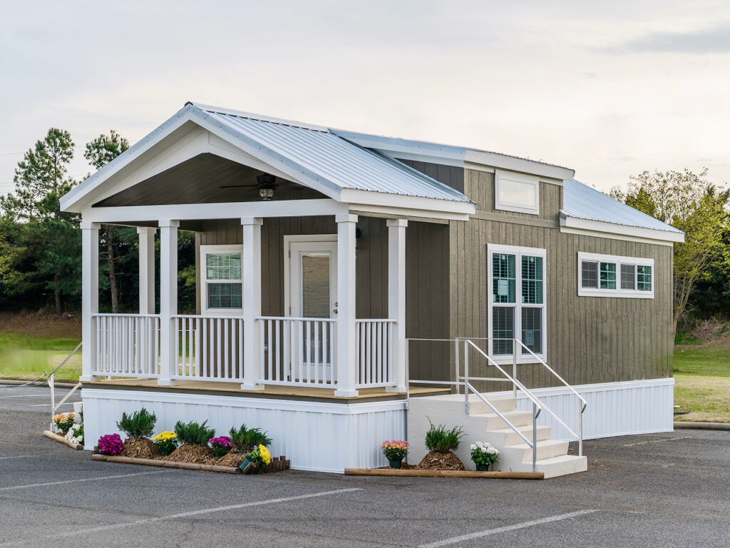 A small, modern tiny house with a gray exterior, white trim, and a metal roof is situated in a parking lot. Bright flowers decorate the area around the porch.