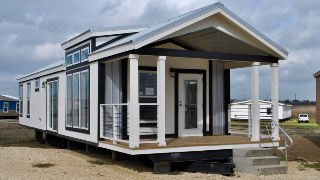 A modern tiny house with large windows, a small porch with railings, and white and black trim. It sits on a gravel lot under a cloudy sky.