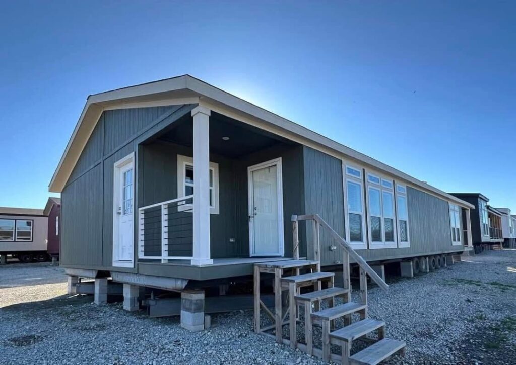 A green manufactured home with a small porch and white door is elevated on blocks, set against a clear blue sky. Steps lead up to the entrance.