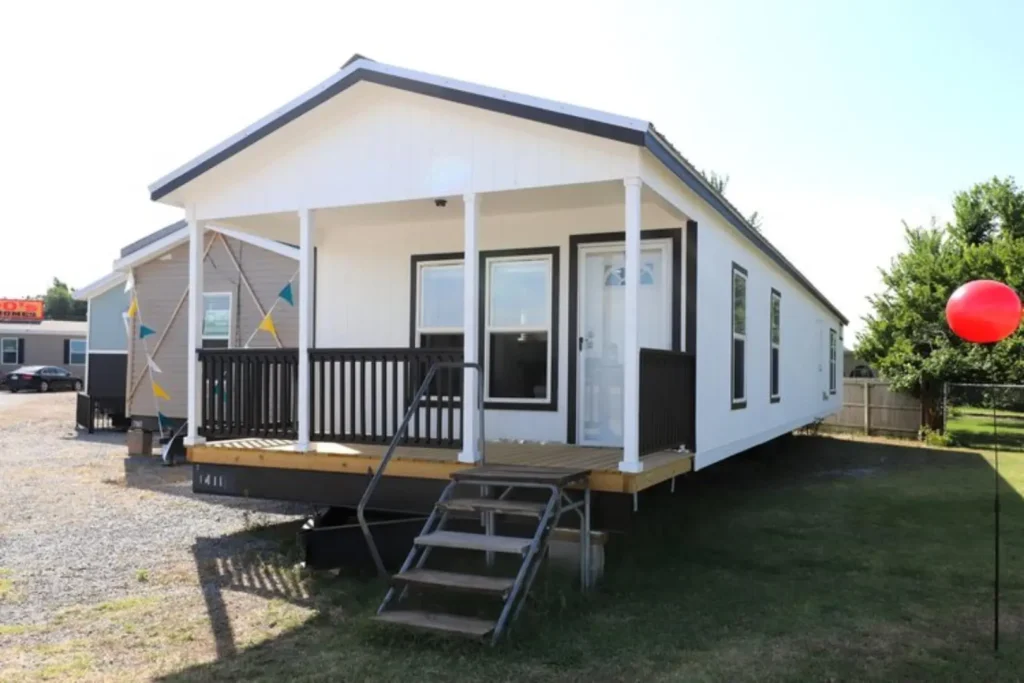 A modern white and black mobile home with a small porch and steps is surrounded by grass. Colorful flags hang, and a red balloon floats nearby.