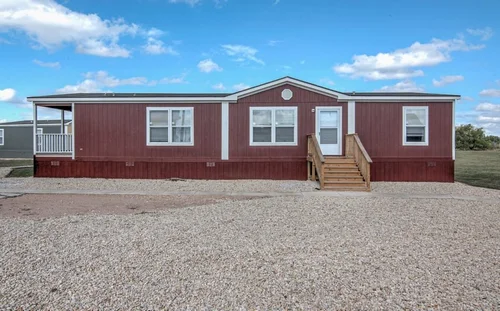A red mobile home with a porch sits on a gravel lot under a blue sky with scattered clouds. The image conveys a sense of simplicity and tranquility.