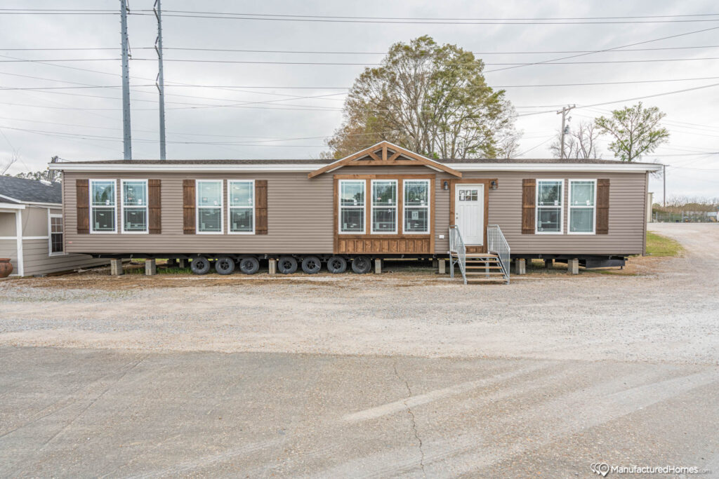Long brown mobile home with large windows and a small front porch sits on a gravel lot. Overcast sky and sparse trees in the background.