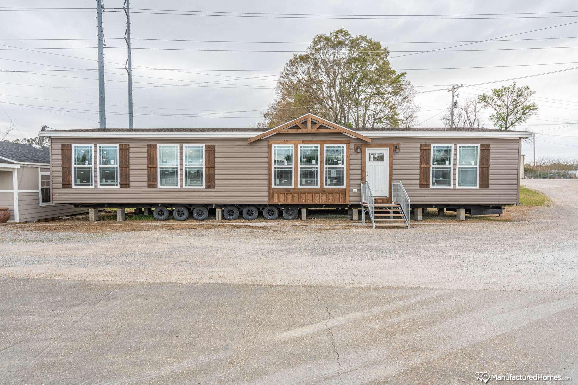 Long brown mobile home with large windows and a small front porch sits on a gravel lot. Overcast sky and sparse trees in the background.