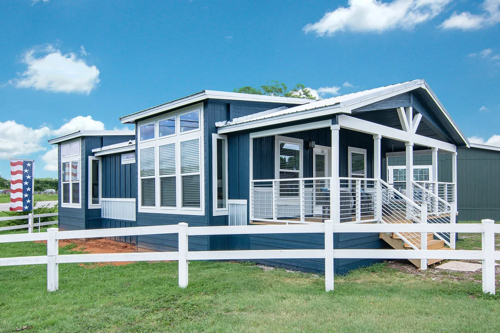 Single-story blue house with white trim, large windows, and a porch with cable railing. It is surrounded by a white fence on a grassy lawn. Bright, sunny day.