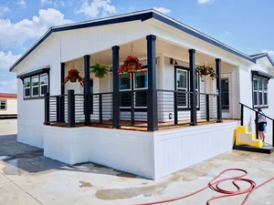 Spacious white modular home with a covered porch, black railings, hanging plants, and yellow steps. A red hose is coiled on the concrete driveway.