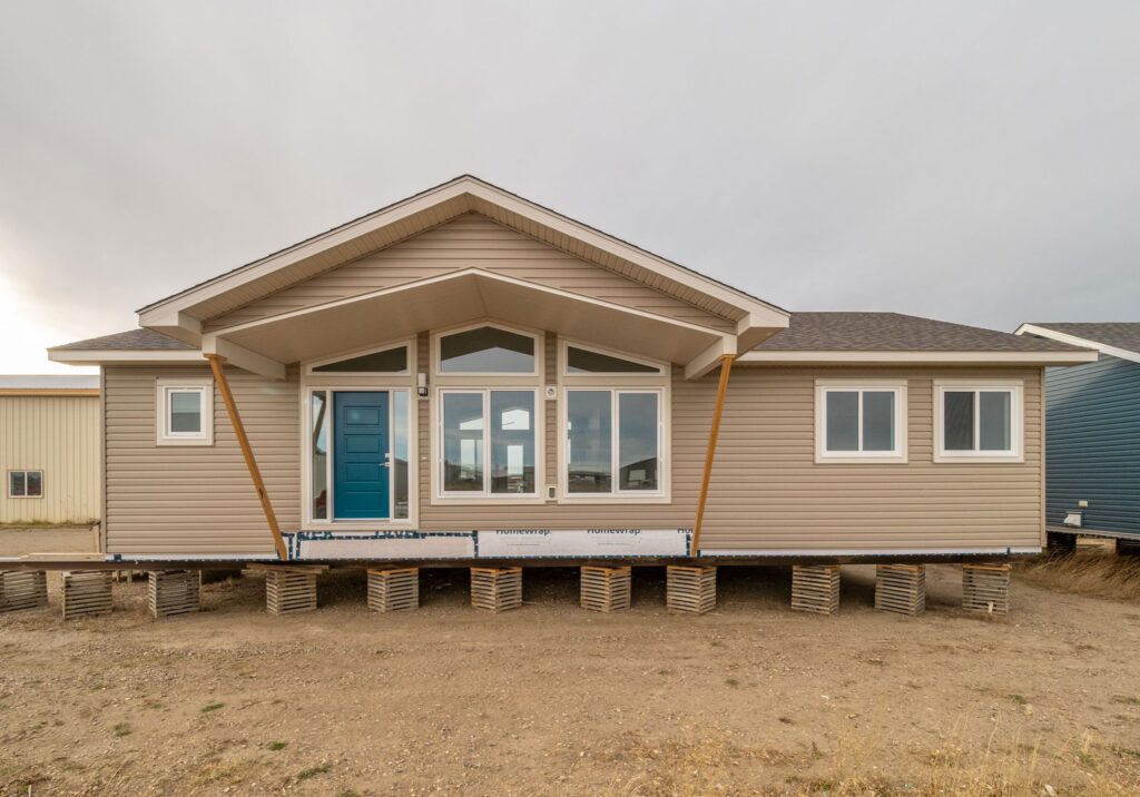 A tan modular home on raised blocks, with a peaked roof and a prominent blue front door. Large windows reflect the overcast sky, conveying a calm, neutral tone.