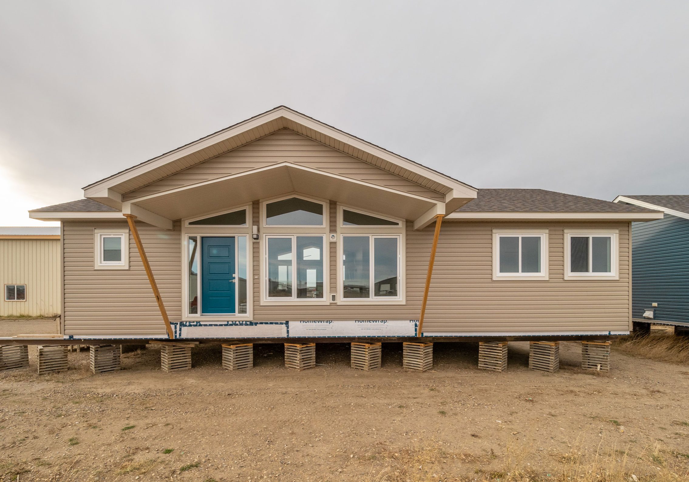 A tan modular home on raised blocks, with a peaked roof and a prominent blue front door. Large windows reflect the overcast sky, conveying a calm, neutral tone.