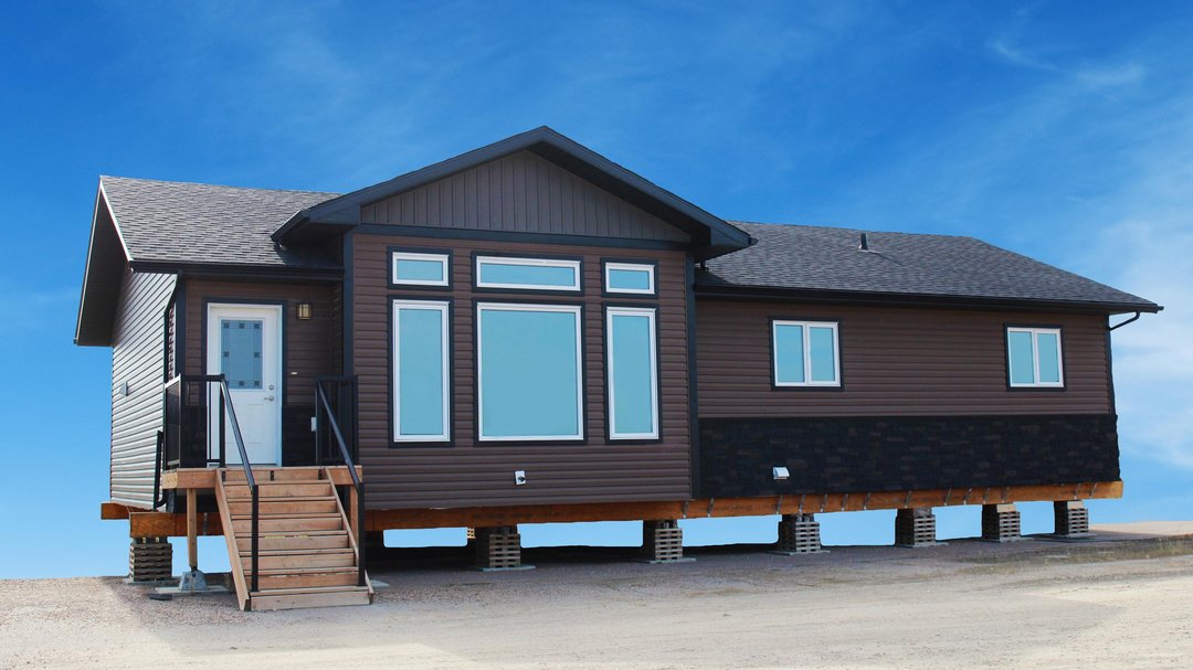 A modern, brown modular home on raised foundations, set against a clear blue sky. Features large windows, a black accents, and a front staircase.