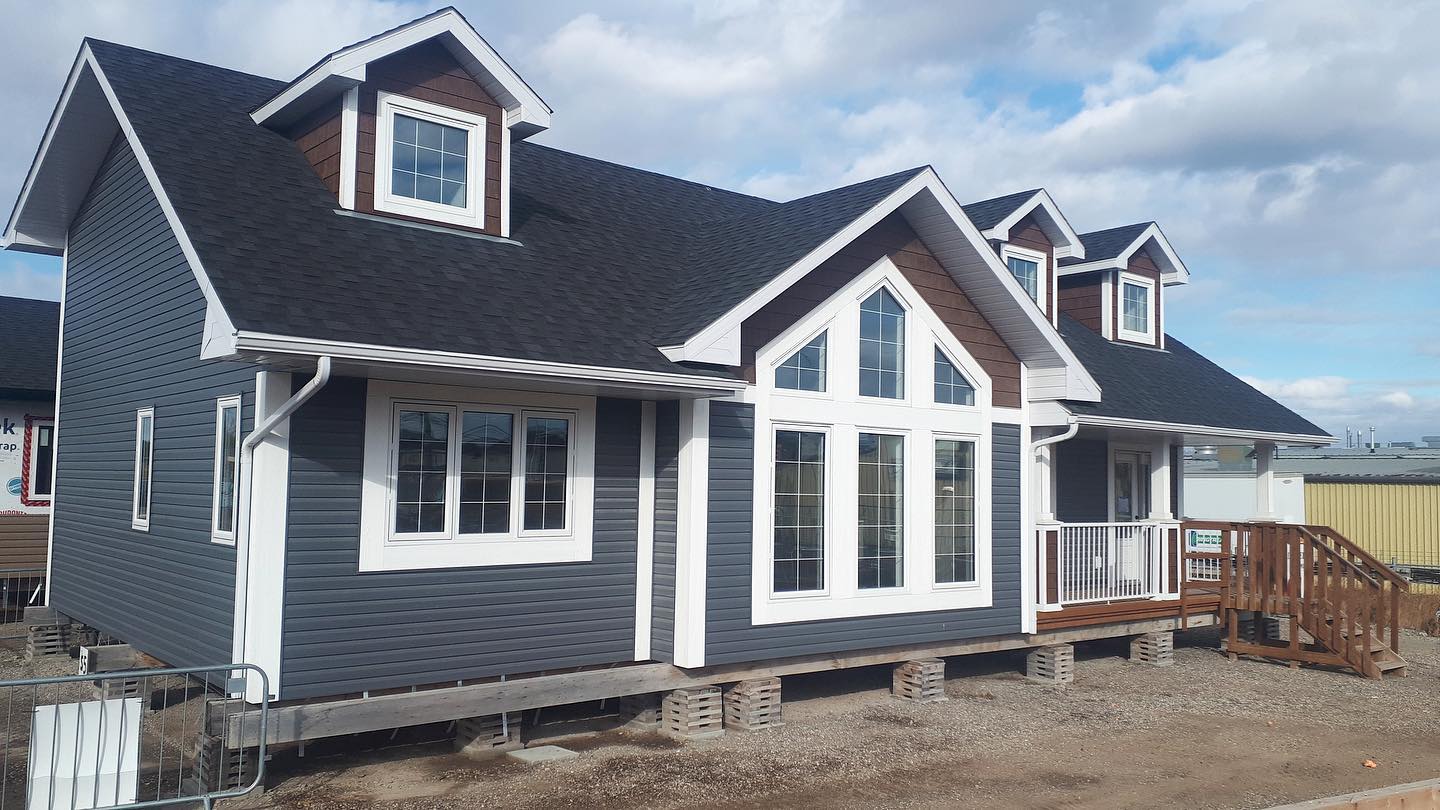 A modern, blue-gray house with white trim and dark roof sits elevated on concrete blocks. It has large windows and a wooden porch under a cloudy sky.