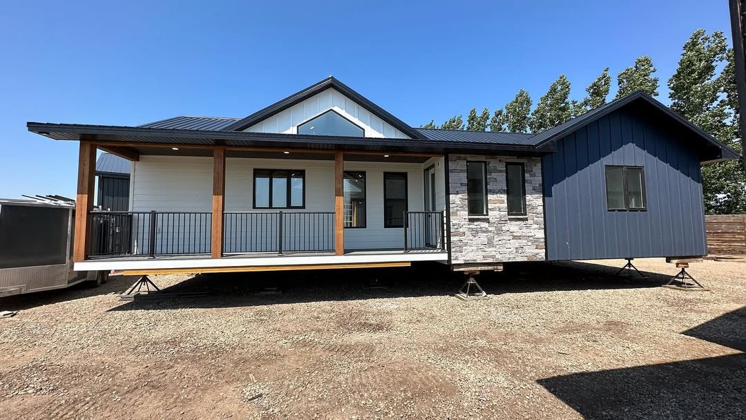 A modern prefab home with a wide porch, featuring wooden pillars, white siding, and stone accents. The house rests on temporary supports, under a clear blue sky.