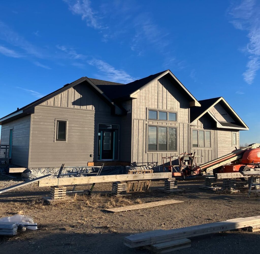 Grey modern house under construction against a blue sky, with scattered building materials and equipment in the foreground.