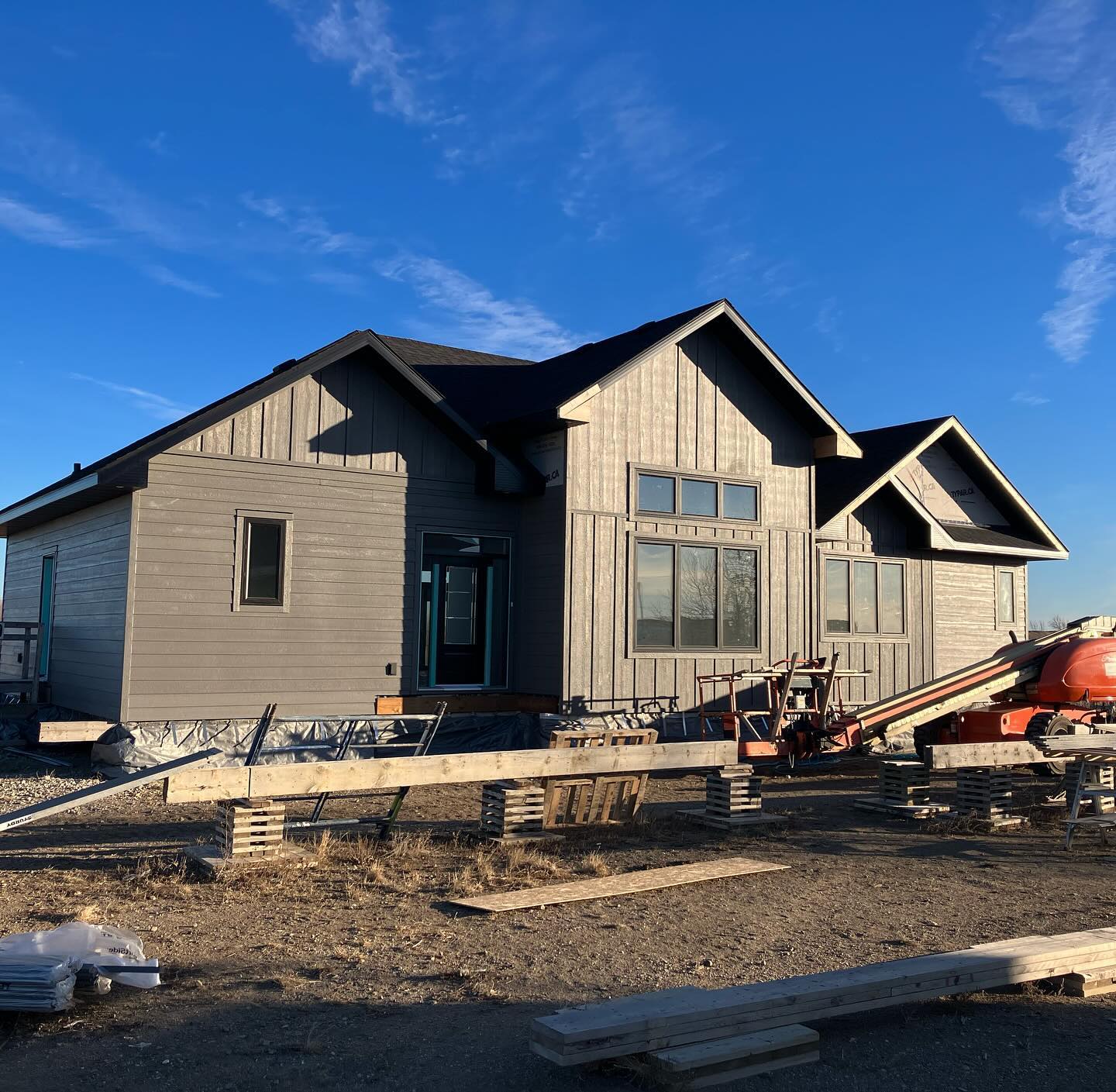 Grey modern house under construction against a blue sky, with scattered building materials and equipment in the foreground.