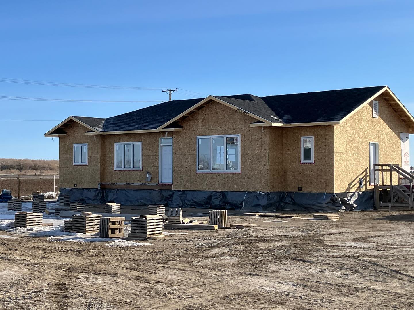 Single-story house under construction with unfinished wooden exterior on a dirt lot. Clear blue sky and stacked wooden pallets nearby.