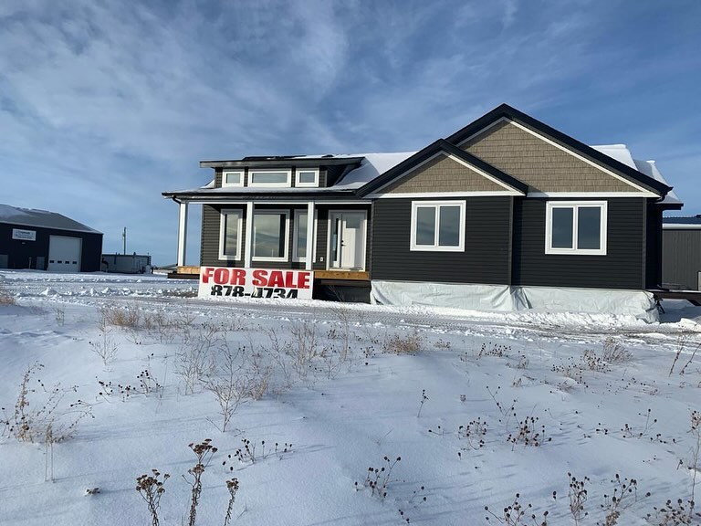 Single-story black and beige house for sale with large front windows and a "For Sale" sign, set in a snowy landscape under a bright blue sky.